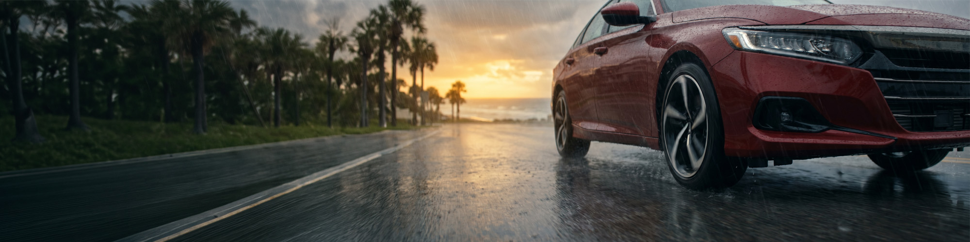 Sedán rojo conduciendo sobre una carretera costera mojada bajo la lluvia al atardecer, destacando el agarre y el rendimiento en condiciones húmedas